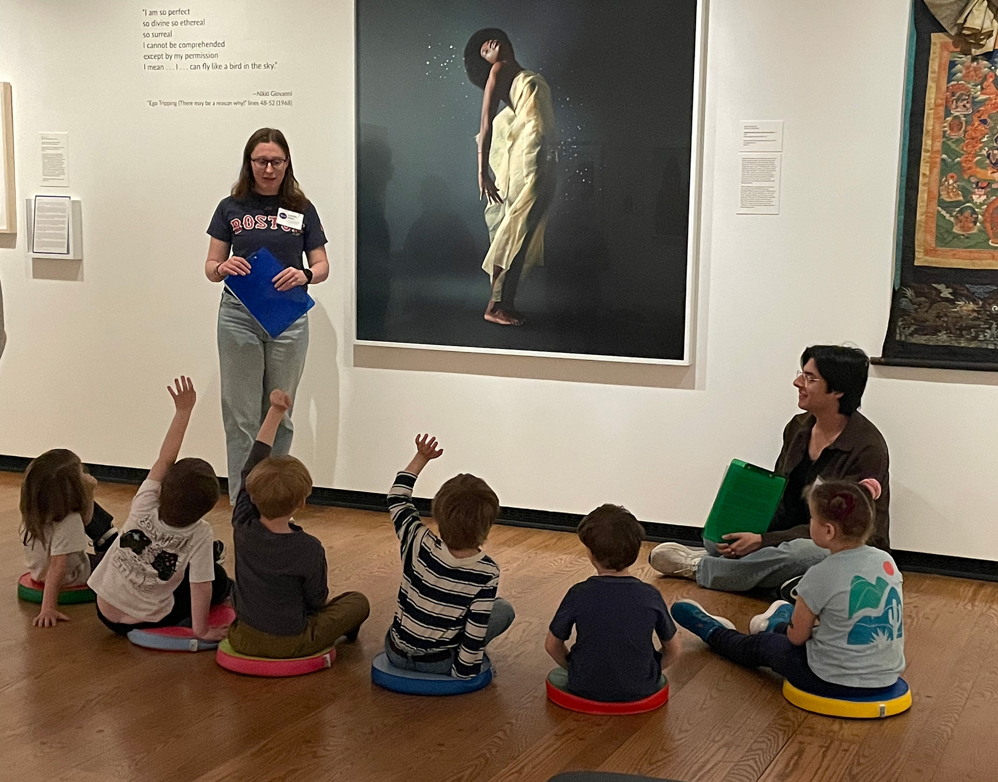 A woman standing in front of a large photograph on a museum wall with children sitting on the floor around her.